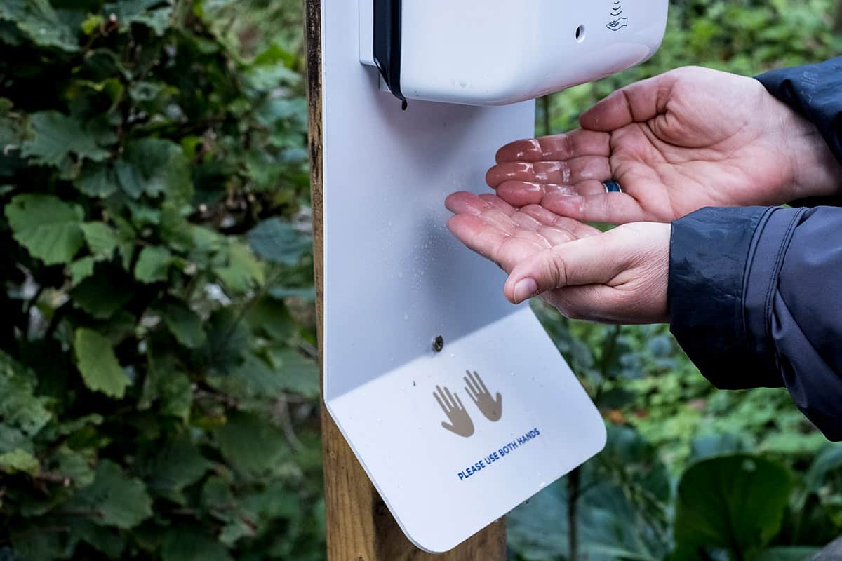 A wall-mounted automatic hand sanitiser dispenser on a wooden post outdoors, a sign beneath it reading Please Use Both Hands, and a pair of hands held underneath the nozzle.