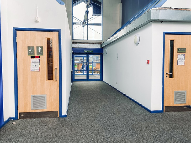 A corridor with a wooden door signed for both a disabled toilet and a ladies toilet on the same sign, and a meeting room door opposite.