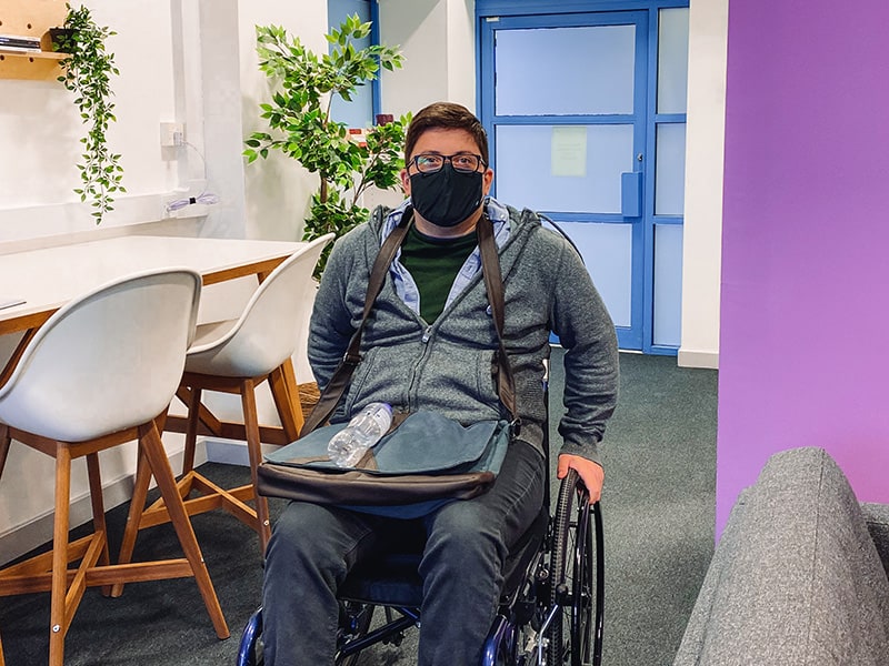 Dean sitting in his wheelchair in an office reception, facing the camera, with a bag across his lap and blue double doors behind him.