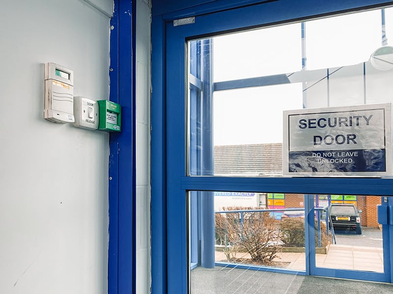 A green door release button mounted high on a wall, well above the reach of someone seated in a wheelchair.