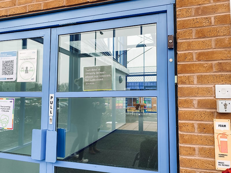 A locked glass entrance door with a notice telling visitors to use the intercom, and the intercom panel mounted high on the brick wall beside it.