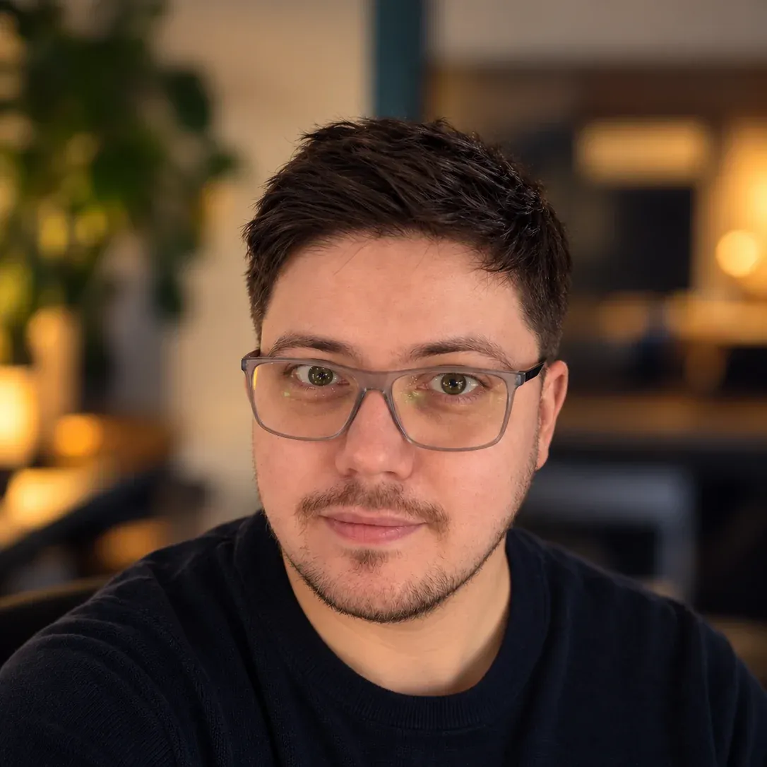 Dean, facing the camera with a slight smile, wearing grey-framed glasses and a dark navy jumper, in a warmly lit cafe with soft lights and a plant blurred behind him.